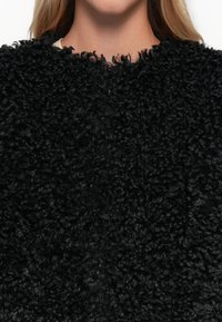 Μαύρος, σγουρός πλεκτός πουλόβερ με στρογγυλή λαιμόκοψη. Το ύφασμα φαίνεται αφράτο και παχύ, δημιουργώντας μια απαλή, χνουδωτή αίσθηση. Χωρίς ορατές μεταλλικές λεπτομέρειες ή σχέδια.