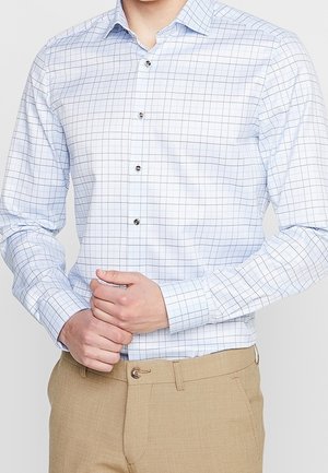 Man wearing a white button-up shirt with light blue grid pattern and beige pants, adjusting his shirt cuff with both hands.