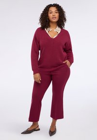 Pull en tricot bordeaux avec un décolleté en V prononcé sur une chemise beige à col, associé à un pantalon évasé bordeaux et des ballerines marron à bout pointu.