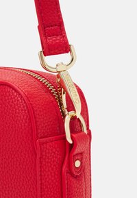Borsa in pelle rossa texture con hardware dorato, dotata di chiusura a zip e tracolla regolabile. Dettagli degni di nota includono un tirazip personalizzato.