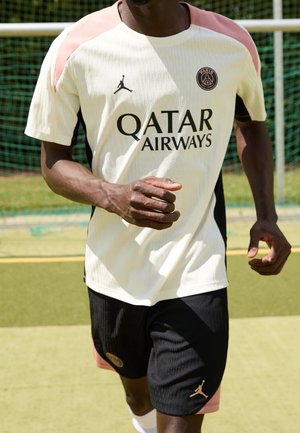 Mann im PSG-Fußballtrikot läuft auf einem Freiplatz, trägt weißes Trikot mit pinken Akzenten und schwarze Shorts, Nahaufnahme des Oberkörpers und der Arme.