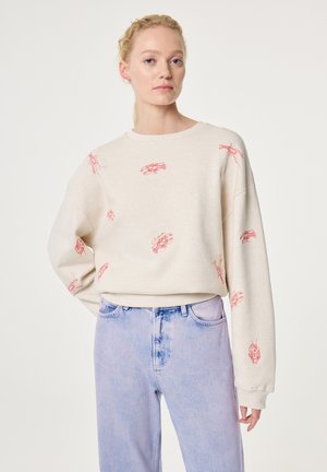 Vrouw met lichtbeige sweatshirt met rode kreeftenprint en lichtblauwe high-waist jeans, staand met één hand op de heup tegen een witte achtergrond.