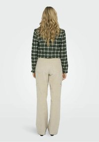 Femme blonde vue de dos portant une veste à carreaux verte et un pantalon large beige sur un fond clair uni.