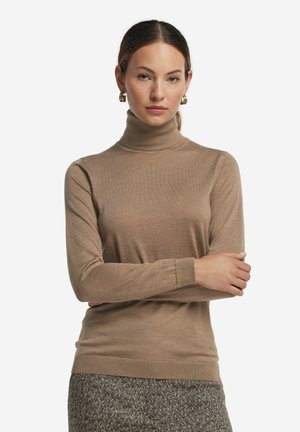 Kobieta ubrana w beżowy sweter z golfem i wzorzyste spodnie, z założonymi rękami i złotymi kolczykami kołami, na jasnym, jednolitym tle.