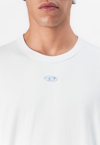 Man wearing plain white crew-neck T-shirt with a small blue oval logo centered near the neckline.