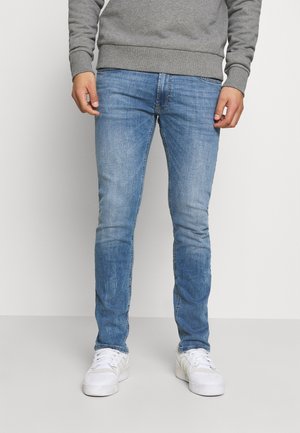 Ein Mann trägt hellblaue Slim-Fit-Jeans, weiße Turnschuhe und einen grauen Pullover und steht vor einem einfarbigen hellen Hintergrund.