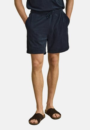 Man wearing navy drawstring shorts and dark brown slip-on sandals, standing with hands in pockets against plain background.