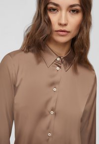 Blusa a maniche lunghe di colore marrone uniforme, realizzata in tessuto morbido con colletto a punta e bottoni perlati sul davanti.