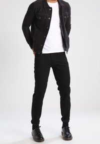 Chaqueta de mezclilla negra con dos bolsillos frontales, llevada sobre una camiseta blanca. Unos pantalones negros y zapatos negros completan el conjunto.