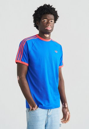 Blauw katoenen T-shirt met rode accenten en drie rode strepen op de schouders. Ronde hals, korte mouwen en een logo op de borst.