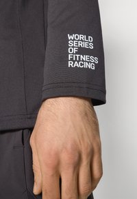 Svart, elastisk treningsskjorte med lange ermer som har hvit tekst som står "WORLD SERIES OF FITNESS RACING." Glatt tekstur, tettsittende design.
