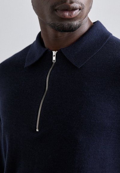 Marineblaues Polohemd mit Reißverschluss und spitzem Kragen sowie einem silbernen Reißverschluss, der ein kleines 'B'-Logo zeigt. Glatte Stricktextur. Nahaufnahme.