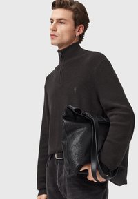 Jonge man in een zwarte kwart-zip trui en donkere jeans, met een grote zwarte leren tote bag tegen een effen achtergrond.