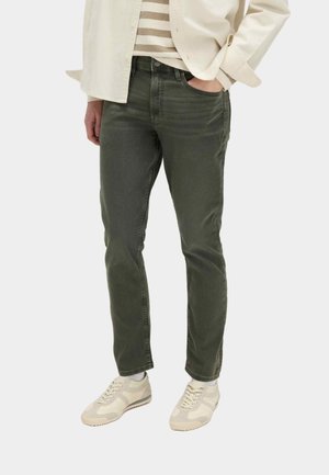 Persona che indossa pantaloni slim fit verde scuro, sneakers beige e bianche, una camicia a righe e una giacca beige chiaro con una mano in tasca.