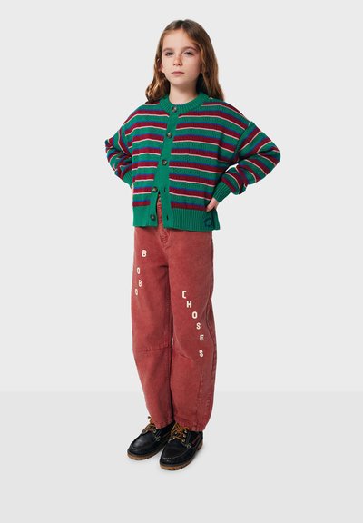 Cardigan rayé vert et rouge avec des boutons, associé à un pantalon couleur rouille avec du texte blanc. Chaussures noires avec des accents marron.