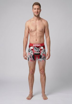 Jonge man staat blootsvoets en draagt rode boxershorts met dollarbiljet- en haaienbekprint tegen een effen grijze achtergrond.