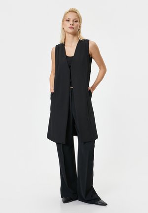 Black sleeveless vest with a long, open design, made of smooth fabric. It features side pockets and is paired with wide-leg black trousers.