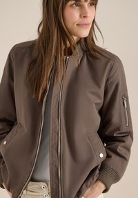 Femme portant une veste bomber marron zippée avec une poche zippée sur la manche et les mains dans les poches latérales, regardant vers la droite