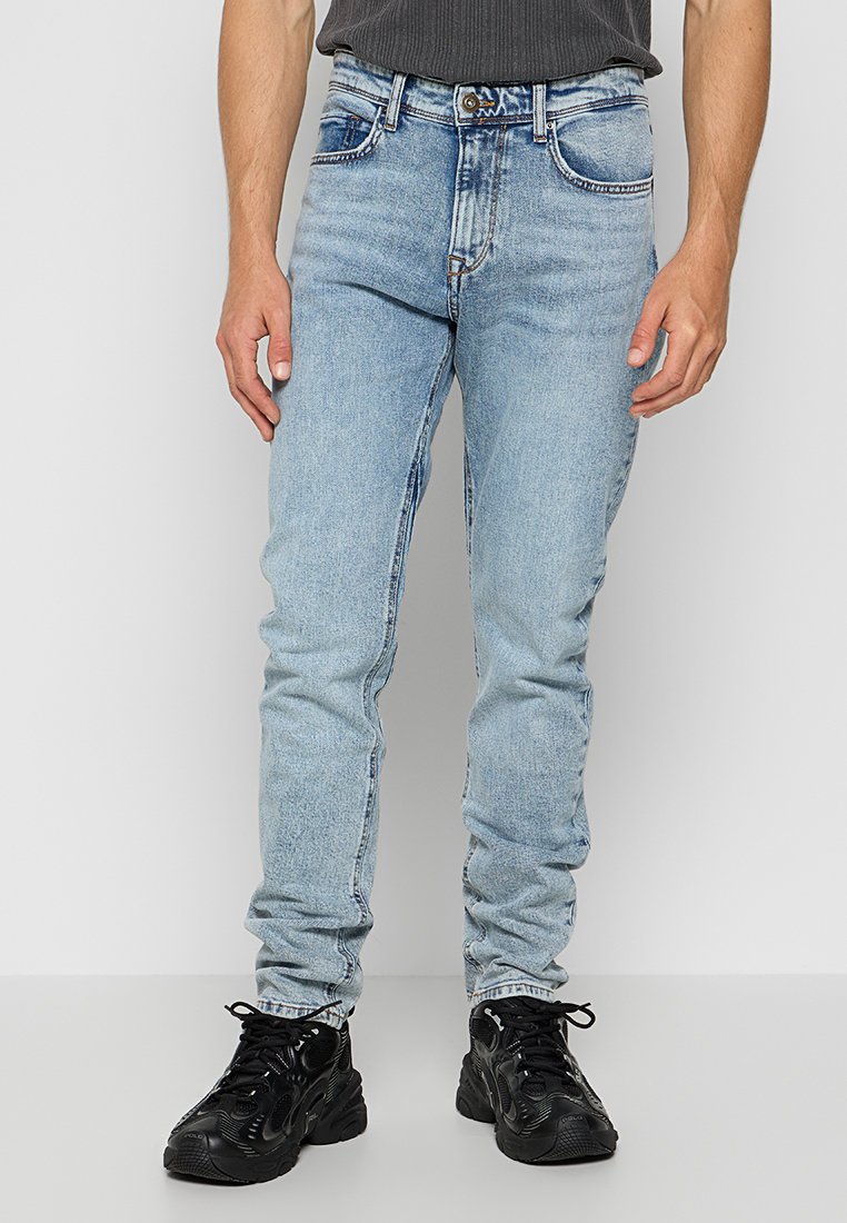 Mann trägt hellblaue Slim-Fit-Jeans und schwarze Schnürschuhe und steht vor einem schlichten weißen Hintergrund.