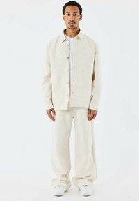 Beige texturerad jacka med knappar, lager på en vit skjorta, som är kombinerad med matchande vida byxor och vita sneakers.