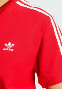 Rød bomulds T-shirt med hvide skulderstriber, med et lille hvidt Adidas-logo på venstre side af brystet. Korte ærmer.