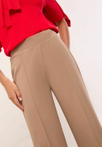 Beige wide-leg trousers with textured fabric and vertical seams, paired with a red blouse featuring a tied neck and rolled sleeves.