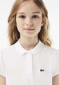 Biała koszulka polo z krótkimi rękawami, falbaniastym kołnierzykiem i listwą guzikową; posiada zielone logo krokodyla na lewej piersi.