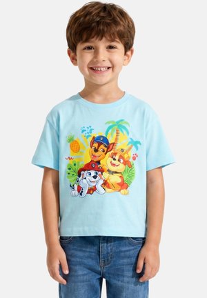 Garçon souriant portant une chemise bleu clair avec des chiots dessinés colorés et des éléments tropicaux, debout devant un fond uni.