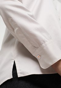 Camicia button-up beige chiaro con maniche lunghe, caratterizzata da una texture liscia, polsini angolati e una fessura laterale all'orlo.