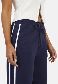 Navy sweatpants met witte zijstrepen, voorzien van een trekkoord in de taille en een zachte katoenstructuur. Een close-up toont de tailleband en de stof.