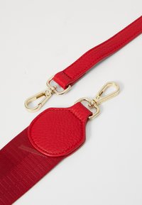 Punane nahkrihm tekstuurse viimistlusega, millel on kuldsete detailide ja ümmargune kinnituskohte. Sujuv kangasosa on integreeritud.