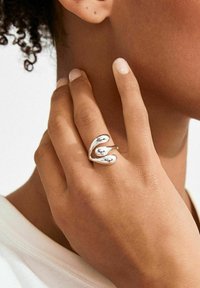 Hõberõngas, millel on kolm kaardus, esile tõstetud osa, sileda ja läikiva viimistlusega. Käsi on kaela lähedal, näidates tekstuurse nahaga.