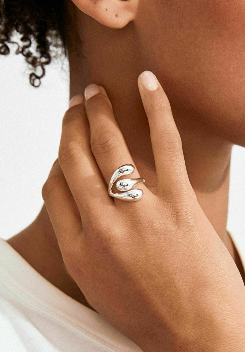 Hõberõngas, millel on kolm kaardus, esile tõstetud osa, sileda ja läikiva viimistlusega. Käsi on kaela lähedal, näidates tekstuurse nahaga.