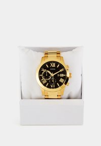 Montre en acier inoxydable doré avec cadran noir, index en chiffres romains, fonctionnalités chronographe et affichage de la date. Présentée sur un coussin blanc dans une boîte.