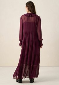 Longue robe bordeaux en tissu transparent. Caractérisée par un col à volants, des manches longues avec poignets élastiques, et une jupe à étages.