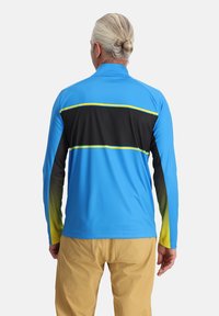 Giacca atletica blu con una striscia orizzontale nera, caratterizzata da accenti gialli e un design sfumato sulle maniche. Texture liscia.