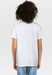 Dijete s kovrčavom kosom nosi jednostavnu bijelu majicu s kratkim rukavima i svijetloplave traperice, okrenuto leđima prema jednostavnoj pozadini.