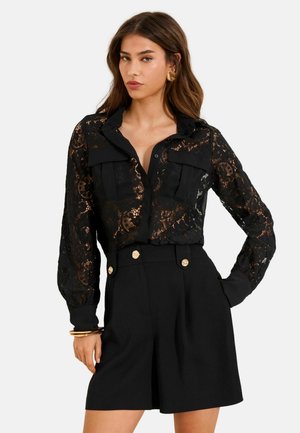 Kobieta nosząca czarną koronkową bluzkę z długim rękawem, z przednimi kieszeniami oraz czarne szorty z wysokim stanem i złotymi guzikami.