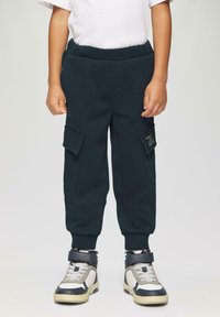 Dunkelblaue Baumwoll-Cargohosen mit elastischem Bund, zwei Seitentaschen und sichtbaren Nähten. Kombiniert mit hohen Sneakers in neutralen Tönen.