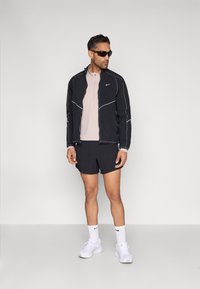 Črna Nike jakna z zadrgo, bele šive, svetlo rožnata srajca z ovratnikom, črne kratke hlače, beli supergi in temna sončna očala.