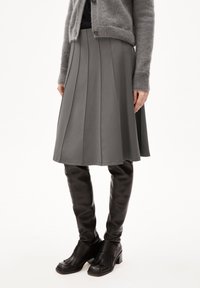 Kvinna klädd i en grå plisserad kjol, grå cardigan, svarta knähöga läderstövlar med klack på en vit bakgrund.