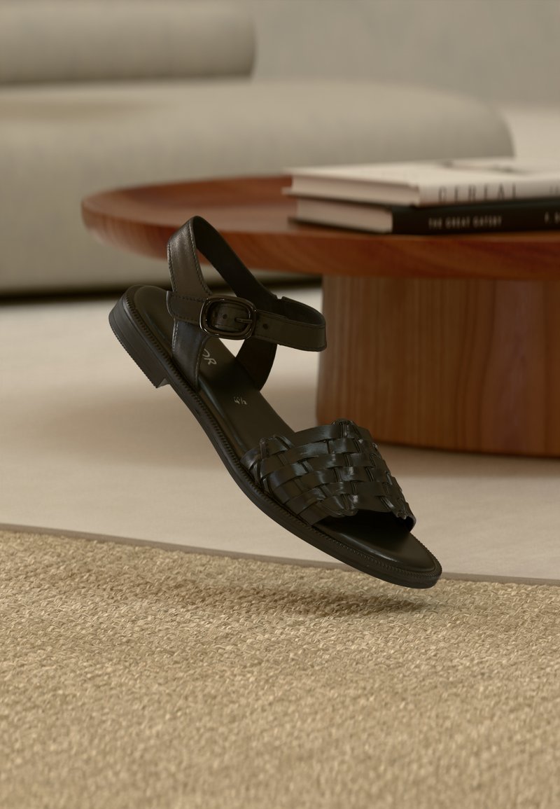 Μαύρο δερμάτινο σανδάλι με πλεκτούς ιμάντες, ρυθμιζόμενο κούμπωμα στον αστράγαλο, επίπεδη σόλα και ανοικτή σχεδίαση στο δάχτυλο, τοποθετημένο πάνω σε υφασμάτινο χαλί με υφή.