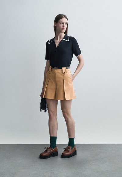 Γυναίκα που φοράει μαύρο τοπ με κοντά μανίκια και λευκή επένδυση, φούστα με πτυχές σε ταμπέ απόχρωση, πράσινες κάλτσες, καφέ παπούτσια με πάτο και κρατάει μία μικρή μαύρη τσάντα χειρός.