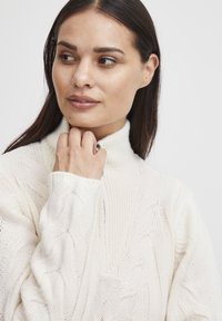 Bílý svetr s pleteným vzorem a vysokým límcem. Měkká textura a detailní vzor po celém svršku. Modelka se jemně dotýká krku, má dlouhé tmavé vlasy.