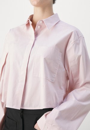 Osoba ubrana w jasnoróżową koszulę na guziki z kieszenią na piersi, w połączeniu z czarnymi spodniami z wysokim stanem, na jednolitym tle.