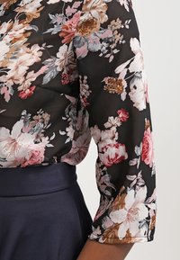 Blusa nera a fiori con maniche a tre quarti, caratterizzata da fiori vivaci in rosa, bianco e beige. Tessuto trasparente con una texture morbida.
