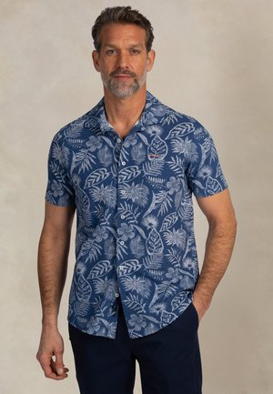 Man met kort donker haar en baard, gekleed in een blauw overhemd met korte mouwen en witte bloemenpatronen, staand met één hand in de zak.