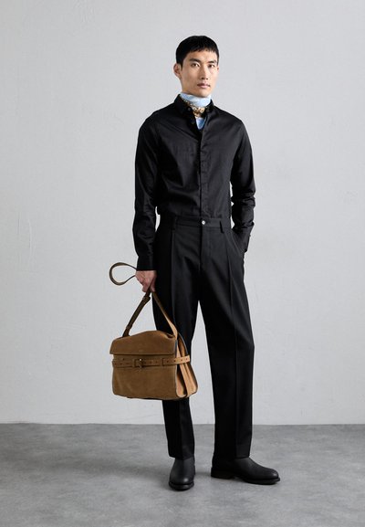 Cămașă neagră cu nasturi, pantaloni eleganți și cizme până la gleznă. Ținând o geantă din suede maro cu clapetă și detalii cu cataramă. Textură netedă.