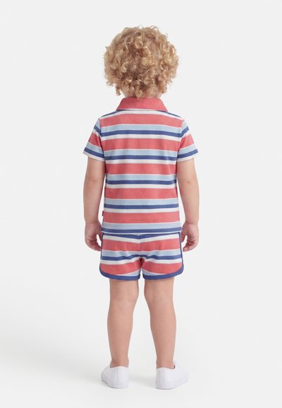Tout-petit aux cheveux bouclés portant un t-shirt à manches courtes rayé rouge, blanc et bleu et un short assorti, debout de dos sur un fond blanc.