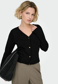 Ženska s kratkimi blond lasmi nosi črno zgornjo s sponkami v obliki zlatih src, bež hlače, zlato nakit in nosi črno mehko usnjeno torbico.